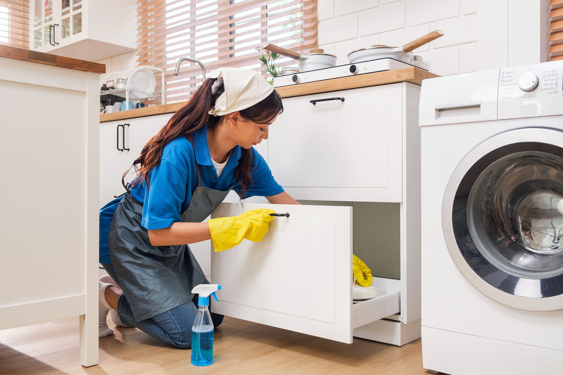 Professional maid in uniform, gloves, and apron cleans a modern kitchen. She organizes cabinets and uses eco-friendly spray for hygiene. Home cleaning service ensures a tidy, fresh, and spotless space